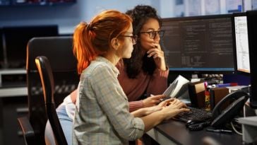 Two engineers look at a computer monitor to review the work being done on an AI project. 