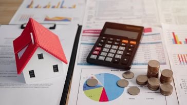 A calculator, stacks of coins and a small model of a house sit on top of financial documents filled with charts and graphs.