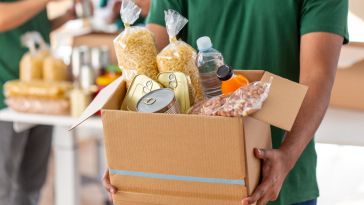 Person carrying cardboard box filled with shelf-stable food items.