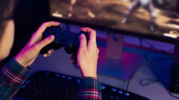 A close up of a pair of hands holding a game controller in front of a PC monitor.