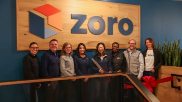  Eight Zoro leaders stand with oversized scissors in front of a logo wall, ready to cut the red ribbon in front of them for their office grand opening. 