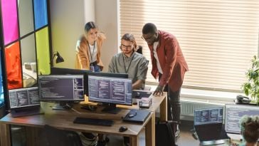 Three technologists analyze data together on a computer monitor screen in an office