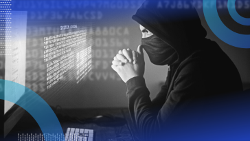 A stereotypical hacker sitting in front of a computer wearing a black hoodie and face mask, hands clasped in front of his face in concentration as he stares at holographic code that’s hovering off the screen.