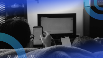 A view from behind of two people sitting on a couch, watching TV while also being on their smartphones.