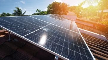 Solar panels sit on top of a roof with trees and sun shining in the background.