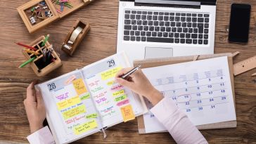 Hands write notes in a daily planner and a paper calendar on a busy work desk.