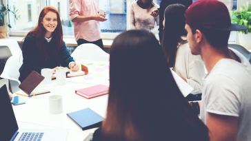 A group of young professionals collaborating in a brightly lit office space. One woman smiles while writing in a notebook at a table surrounded by laptops, notebooks, coffee mugs and sticky notes. Others are standing and chatting in the background near large windows.