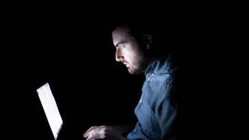 A man sitting in a dark room and staring at a computer screen.