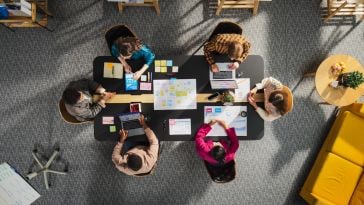 Overhead shot of five people collaborating around a black meeting table in an office. They are looking at laptops, tablets, and sticky notes on the table.