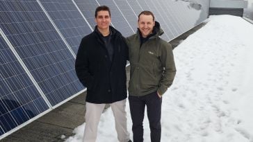 Two men stand next to solar panels in the snow wearing winter coats and boots and smiling at the camera.