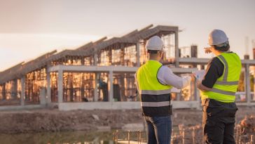 Construction workers in bright green vests and white hard hats standing at a job site are shown.