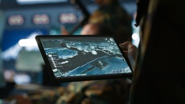 Images of outdoor terrain are displayed on a tablet device being held by a person in a military uniform to represent defense intelligence.