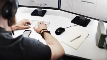 An employee wearing a blackshirt and a headset sits a desk typing on a keyboard in front of two desktop computer monitors.