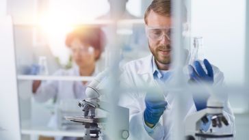 A scientist wearing a white lab coat, goggles and blue gloves is pictured sitting behind a microscope.