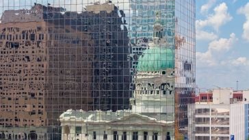 Reflection of a historic dome and buildings on a mirrored skyscraper. The distorted image captures a clear blue sky and clouds, creating a surreal scene.