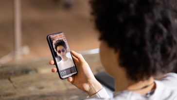 A person holds a smartphone displaying a virtual try-on app, featuring them wearing augmented reality sunglasses. The setting is casual and modern.