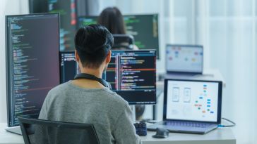 A person sits at a desk with multiple monitors displaying code and diagrams. The setting is a bright office environment, conveying a focused, tech-driven atmosphere.