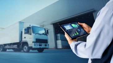 Person holding a tablet with logistics data charts, standing near a white delivery truck outside a warehouse. The scene conveys efficiency in transport management.