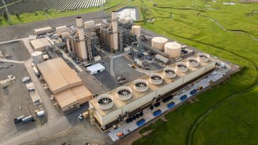 Aerial view of an industrial power plant with cylindrical cooling towers, metal chimneys, and expansive green fields in the background under a clear sky.