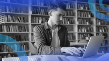 A student works on a laptop next to an open book in a library