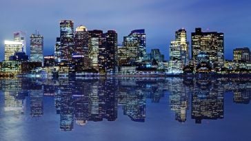 The Boston skyline along the water at night with several skyscrapers lit up and reflected in the water.