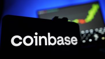 The Coinbase logo is featured on the screen of a smartphone in front of a laptop with a line graph on the monitor.