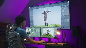 A game developer sits at a desk in a dark room working on animations across three different computer monitors.