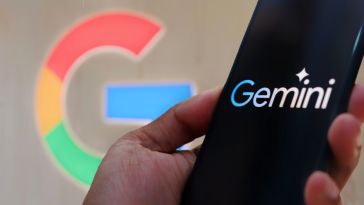 A hand holds a smartphone with the Gemini logo on the screen, and a multi-colored G representing the Google brand is in the background of the image.