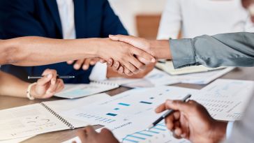 A close up of a handshake over papers spread across a conference room table.