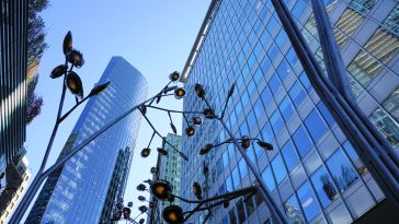 A cluster of modern skyscrapers under a clear blue sky, framed by an artistic metal sculpture with abstract flower shapes in the foreground.