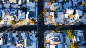 Aerial view of a snowy suburban neighborhood at sunrise, showing a grid of houses with snow-covered roofs and long shadows, creating a tranquil winter scene.