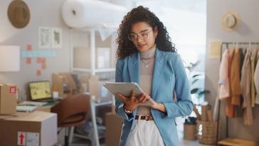 A woman in a blue blazer, standing in a stylish office space, looks at a tablet. Behind her are boxes, a computer, clothes on a rack, and soft lighting.