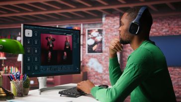 A man in a green sweater and headphones sits at a desk, focused on a computer screen displaying graphic design software. The room has a modern, cozy feel.