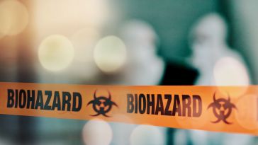 Orange tape featuring the word BIOHAZARD in front of a blurred image of two workers in white protective gear.