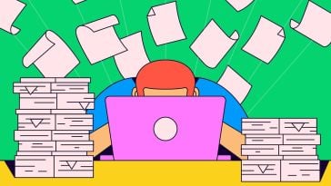 Papers flutter in the background as a worker sits behind a laptop, with stacks of additional paper on either side of them.