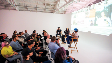 A group attends a VSCO event in a white room with three speakers sitting on director's chairs in front of a projected screen. 