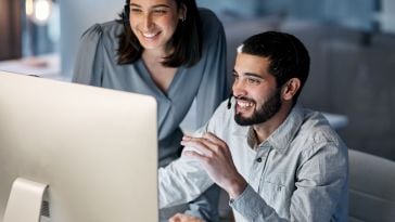 A smiling woman with dark hair stands behind a smiling man with a beard and dark hair, both wearing headsets and looking at a bright computer monitor in a dimly lit office.