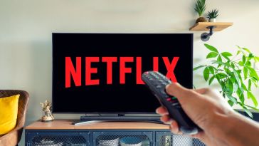 The red and black Netflix logo on a TV in a living with a hand holding up a remote control in front of the screen.