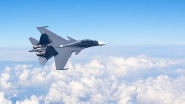 A sleek fighter jet with a pointed nose flies above fluffy white clouds against a clear blue sky, conveying speed and precision in open air.