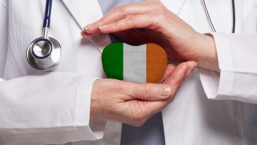 A doctor in a white coat holds a heart-shaped object with the Irish flag. A stethoscope is visible, conveying care and healthcare themes.