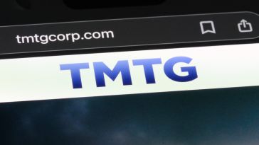 Close-up of a smartphone screen showing the URL "tmtgcorp.com" and a bold blue "TMTG" logo. The background is dark with a cloudy sky.