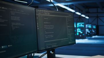 Two computer monitors displaying code in a dimly lit room, with servers in the background, conveying a focused, high-tech environment.