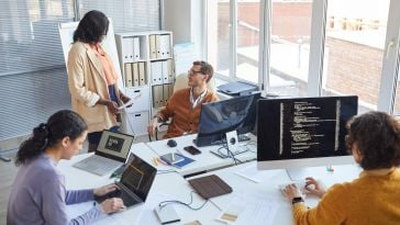 Two software engineers in an office discuss details about a project while two other engineers write code beside them