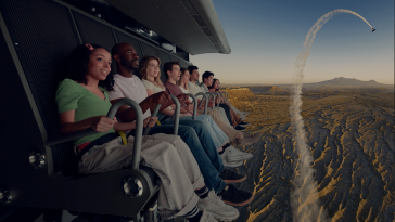 A group of people are sitting in a row on a simulated ride, appearing to fly over a rugged desert landscape. A plane leaves a smoke trail in the sky.