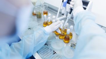 A scientist wearing white gloves and other lab gear uses a pipette in containers of yellow liquid.