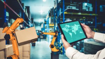 A person holds a tablet displaying a world map in a warehouse. Robotic arms lift boxes, highlighting automation and technology in logistics.