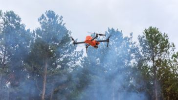 A bright orange drone hovers in front of tall, green pine trees with smoky haze. The scene suggests an outdoor operation in a wooded area.