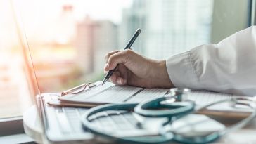 A person in a white coat writes on a clipboard with a pen. A stethoscope rests on the table beside them. A cityscape is visible through the window in the background.