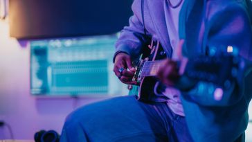 A person playing an electric guitar in a dimly lit room with blue and purple lighting. A computer screen with audio software is visible in the background.