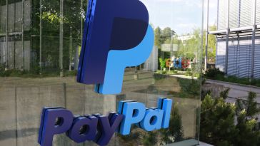 3D PayPal logo on a glass surface, reflecting exterior greenery and a modern building. The blue logo stands out against the lush backdrop.